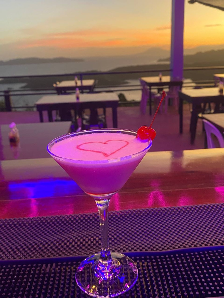 A pink cocktail with a heart design on the neon-lit skybar counter, sunset over the islands behind it
